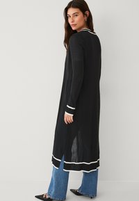 Langer schwarzer Cardigan mit weißen Streifenakzenten, lockerer Passform, hohen seitlichen Schlitzen und leichtem, strukturiertem Stoff.