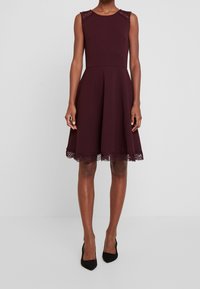 Burgundowa sukienka bez rękawów z koronkowymi akcentami na ramionach, rozkloszowany dół z teksturowanym koronkowym wykończeniem, w zestawieniu z czarnymi szpilkami.