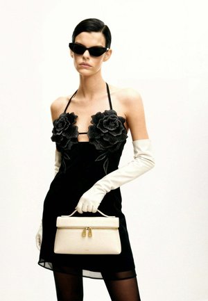 Femme en robe noire à col halter avec de grandes applications florales, gants blancs jusqu'au coude, lunettes de soleil noires, tenant un petit sac à main beige.