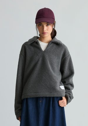 Junge Frau trägt einen dunkelgrauen Fleecepullover, einen blauen Jeansrock, ein weißes Hemd darunter und eine weinrote Baseballkappe vor einfachem Hintergrund.
