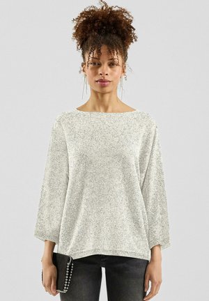 Vrouw met krullend haar draagt een lichtgrijze gebreide trui en zwarte spijkerbroek, houdt een zwarte gestikte clutch vast tegen een effen achtergrond.