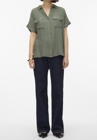 Camicia verde oliva a maniche corte con bottoni e due tasche sul petto. Abbinata a jeans blu scuro a gamba larga e scarpe nere. Tessuto dalla superficie liscia.