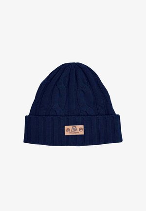 Navy knit beanie, scarf, and gloves with cable patterns; leather logo accent; worn with a light beige wrap coat.