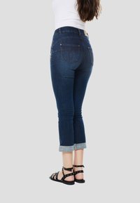 Sötétkék, felhajtott szegélyű, visszavágott farmernadrágok hátsó zsebekkel, fekete, szegecsekkel díszített pántos szandálokkal.