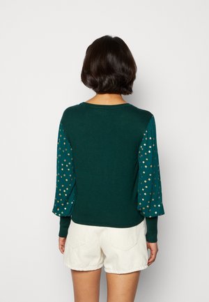 Femme aux cheveux courts et foncés de dos, portant un haut vert foncé avec des manches bouffantes à pois dorés et un short blanc.