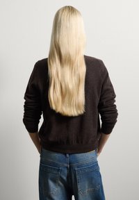 Maglione nero con polsini e orlo a coste, abbinato a jeans blu a vita alta. Lunga chioma bionda e liscia scende lungo la schiena.