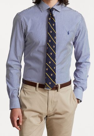 Man draagt een blauw-wit gestreept overhemd, een marineblauwe stropdas met gele diagonale strepen en wapenschilden, beige broek en een bruine riem, handen in de zakken.