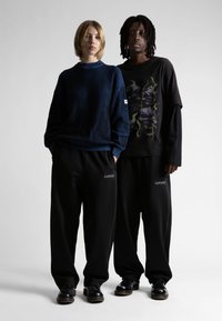 Dva stojna modela, ki nosita ohlapne črne hlače in črne čevlje; eden v modrem puloverju, drugi v črni majici z dolgimi rokavi z grafiko.