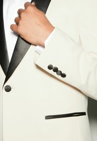 Veste de smoking ivoire avec un revers châle en satin noir, en tissu texturé, et quatre boutons ronds noirs sur le poignet ainsi qu'un détail de poche élégant.