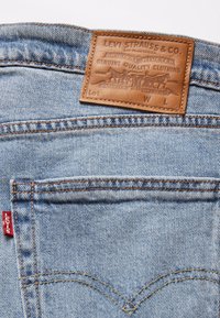 Vaqueros de mezclilla azul claro con una parche de cuero marrón con texto en relieve. Costura clásica en los bolsillos traseros en forma de V, detalle de etiqueta roja.