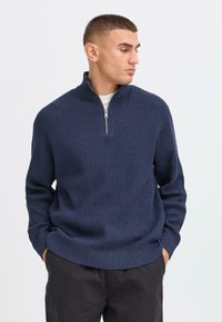 Pull à col zippé navy avec un motif gaufré, poignets et ourlet côtelés, fabriqué en tissu tricoté, porté sur une chemise claire.