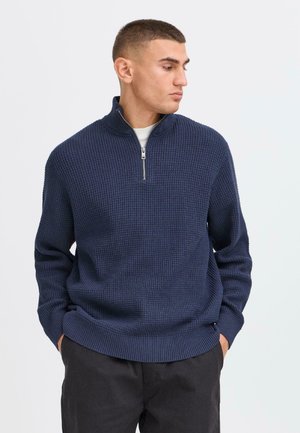 Marinblå zip-tröja med en strukturerad våffelmönster, ribbade ärmslut och nederkant, tillverkad av ett stickat tyg, bärs över en ljus skjorta.