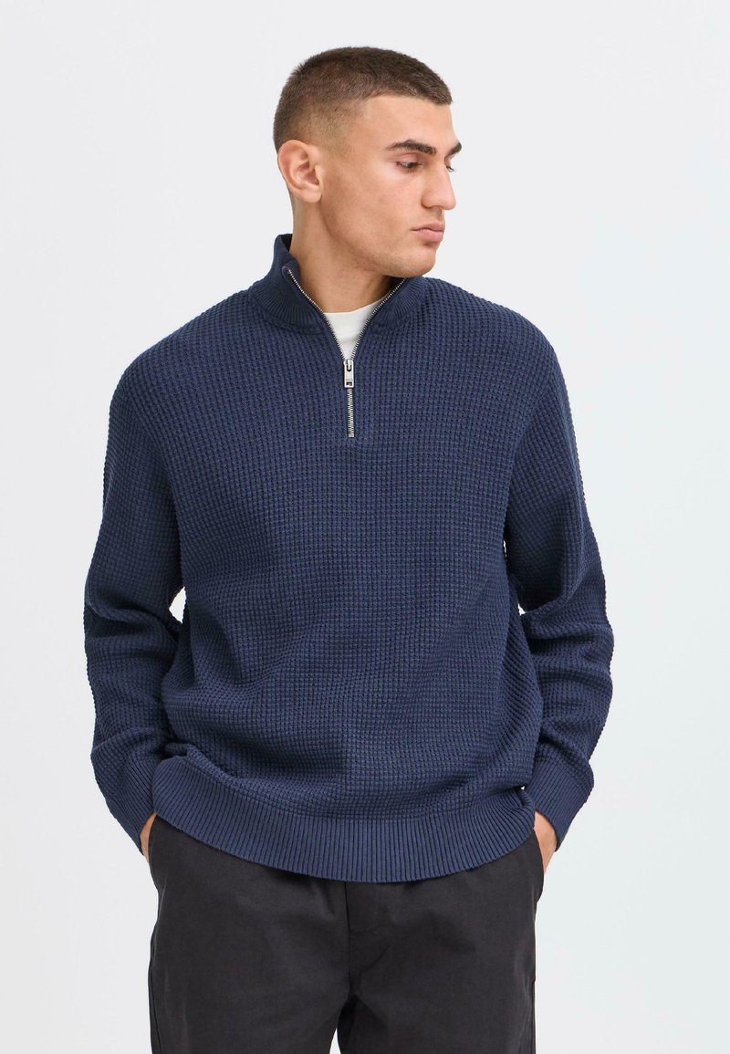 Pull à col zippé navy avec un motif gaufré, poignets et ourlet côtelés, fabriqué en tissu tricoté, porté sur une chemise claire.