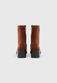 Botine din suede maro, cu vârf pătrat și toc gros negru. Include un sistem de închidere cu fermoar și detalii de cusătură minime.