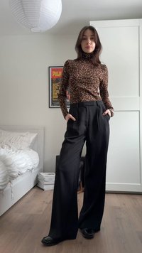 Žena stojící s rukama v kapsách, oblečená v roláku s leopardím vzorem, širokých černých kalhotách a černých botách v prostředí ložnice.