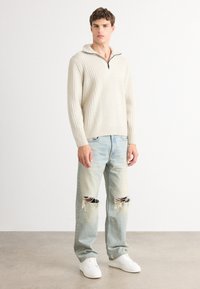 Beige gerippter Strickpullover mit Reißverschlusskragen, kombiniert mit hellblauen, abgewetzten Jeans mit aufgerissenen Knien und weißen Sneakers.
