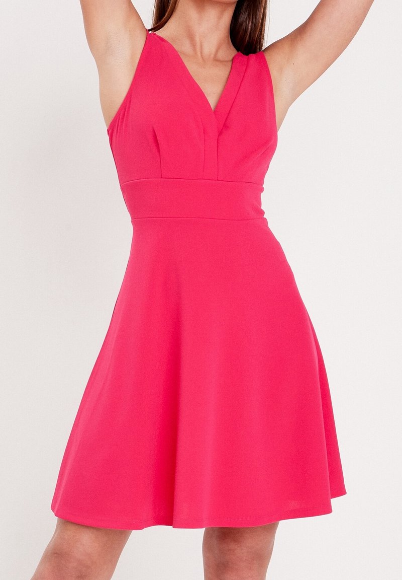 Femme portant une robe sans manches rose vif, avec une taille cintrée et une jupe évasée, bras levés au-dessus de la tête, sur un fond clair uni.