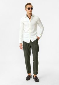 Witte overhemd, olijfgroene, gemusterde broek en zwarte gevlochten instappers. Model draagt een zonnebril. Eenvoudig en op maat gemaakt ontwerp.