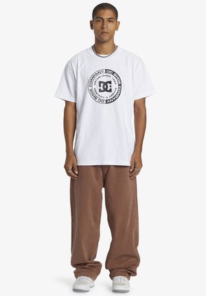 Camiseta blanca de manga corta con un diseño de logo circular negro en el centro; combinada con pantalones marrones holgados y zapatillas blancas.
