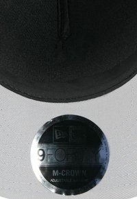 Schwarze verstellbare Kappe mit flacher Krempe, die an der Unterseite glatt grau ist und ein rundes silbernes Etikett mit der Bezeichnung "9FORTY M-CROWN" trägt.