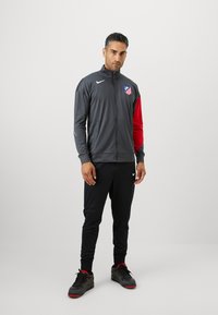 Mørkegrå atletisk jakke med røde ærmer, der har et logo og lynlås. Parret med sorte, tapered bukser og sorte sneakers med røde accenter.