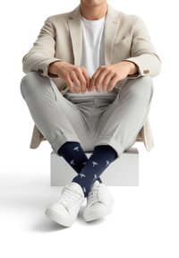 Hombre sentado con las piernas cruzadas, lleva una chaqueta beige, camiseta blanca, pantalones gris claro, calcetines azul marino con patrones de árboles blancos y zapatillas blancas.