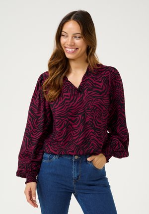 Long-sleeve blouse in fuchsia with black zebra stripes, featuring a V-neck and elasticized cuffs, paired with blue jeans.