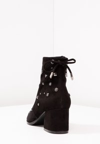 Stivale alla caviglia in suede nero con tacco a blocco, caratterizzato da borchie metalliche e dettagli decorativi. Include una chiusura con lacci e cerniera.