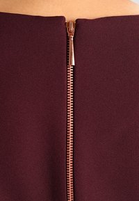 Mörkröd klänning med en synlig, metallisk roséguldfärgad dragkedja längs ryggen, med ett slätt, texturerat tyg och en figursydd silhuett.