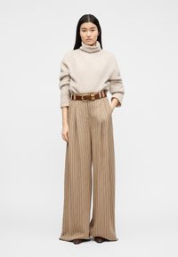 Γυναίκα που φοράει μπεζ πουλόβερ με ζιβάγκο, φαρδιά ριγέ μπεζ παντελόνια με καφέ ζώνη, στέκεται με το ένα χέρι στην τσέπη μπροστά σε λευκό φόντο.