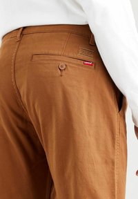 Bruna bomullsshorts med en bakficka med knapp, en Levi's-logotyp och en slät textur. Vit skjorta delvis synlig.