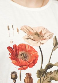T-shirt blanc avec de grands motifs de coquelicot rouges et roses, agrémenté de feuilles vertes et de capsules. Fabriqué dans un tissu doux et lisse.