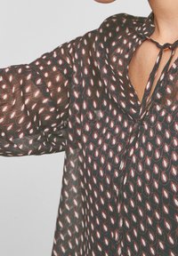Blusa nera trasparente con un motivo circolare in rosso e bianco. Presenta una chiusura con cravatta e un scollo aperto. Tessuto leggero e fluente.