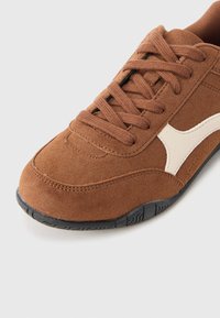 Καφέ σουέντ αθλητικό παπούτσι με μαύρη λαστιχένια σόλα, με σχέδιο με κορδόνια και αντίθετη λευκή δερμάτινη λεπτομέρεια στο πλάι.