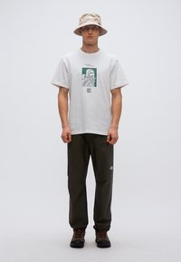 Ανοιχτό γκρι βαμβακερό μπλουζάκι με πράσινο γραφικό σχέδιο, συνδυασμένο με σκούρα πράσινα παντελόνια και μπεζ καπέλο τύπου bucket. Τα παπούτσια είναι σκούρα και ανθεκτικά.