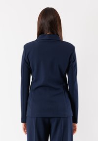 Blazer blu navy realizzato in tessuto liscio, con un design strutturato, maniche lunghe e colletto classico. Il retro presenta una silhouette sartoriale.