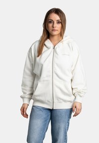 Krémová mikina na zip s velkou kapucí, předními kapsami a žebrovanými manžetami, nošena s světle modrými džíny. Viditelná jemná textura látky.