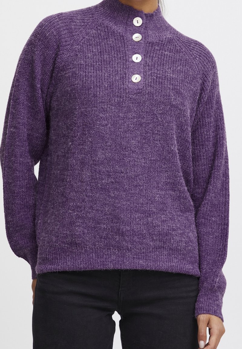 Pull en tricot violet avec col haut et quatre boutons blancs sur le devant, porté avec un pantalon noir.