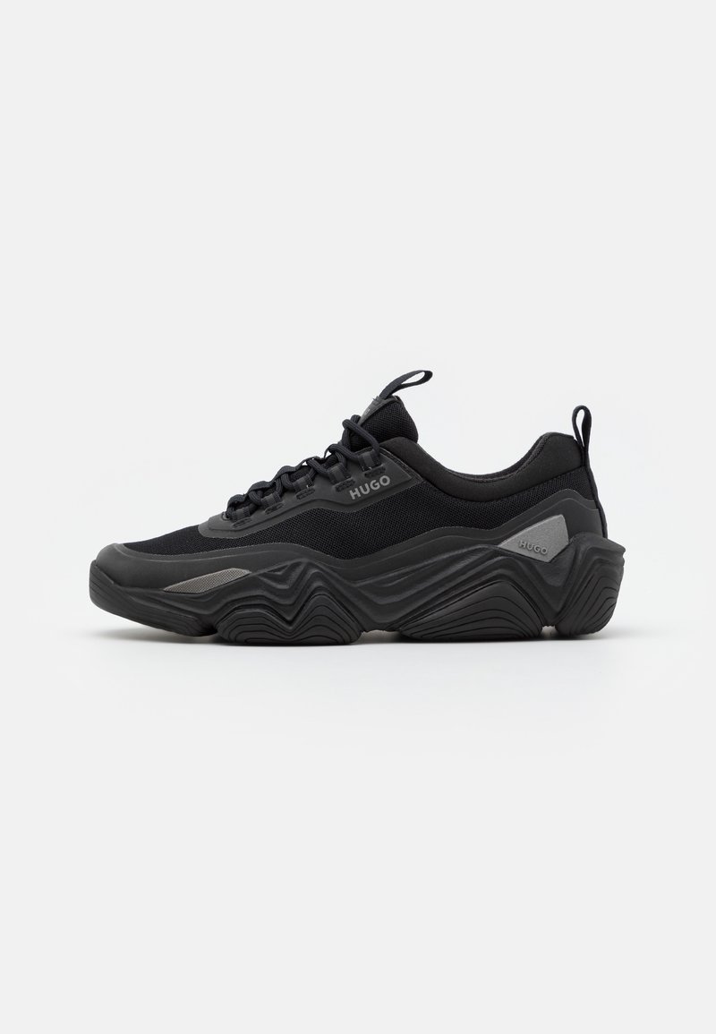 Schwarze Sportschuhe mit einer strukturierten Oberseite, die eine dicke, wellenförmige Sohle und tonale Akzente aufweist. Schnürsenkel und eine Zuglasche runden das Design ab.
