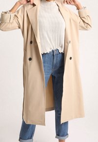 Beige trenjak iz glatkog materijala s dvostrukim kopčanjem, velikim dugmadi i bočnim džepovima, nošen preko svijetlo sivoj rebrastoj majici i plavimfarmericama.