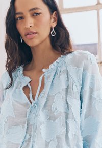 Blusa azzurro chiaro con ricami floreali, tessuto semi-trasparente, scollo rotondo, chiusura con laccetto e dettagli delicati e strutturati su tutta la superficie.