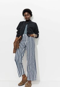 Veste aviateur noire, haut gris, pantalon large rayé bleu marine et blanc, mocassins à imprimé animal, et un sac frangé marron avec des accents dorés.