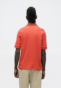 Polokošile s krátkým rukávem z texturované oranžové látky, s klasickým límečkem a volnějším střihem, doplněná béžovými kalhotami.