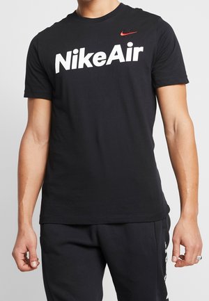 Homme portant un t-shirt noir Nike Air avec un grand texte blanc et un petit logo Nike rouge et noir sur la poitrine, associé à un pantalon noir.