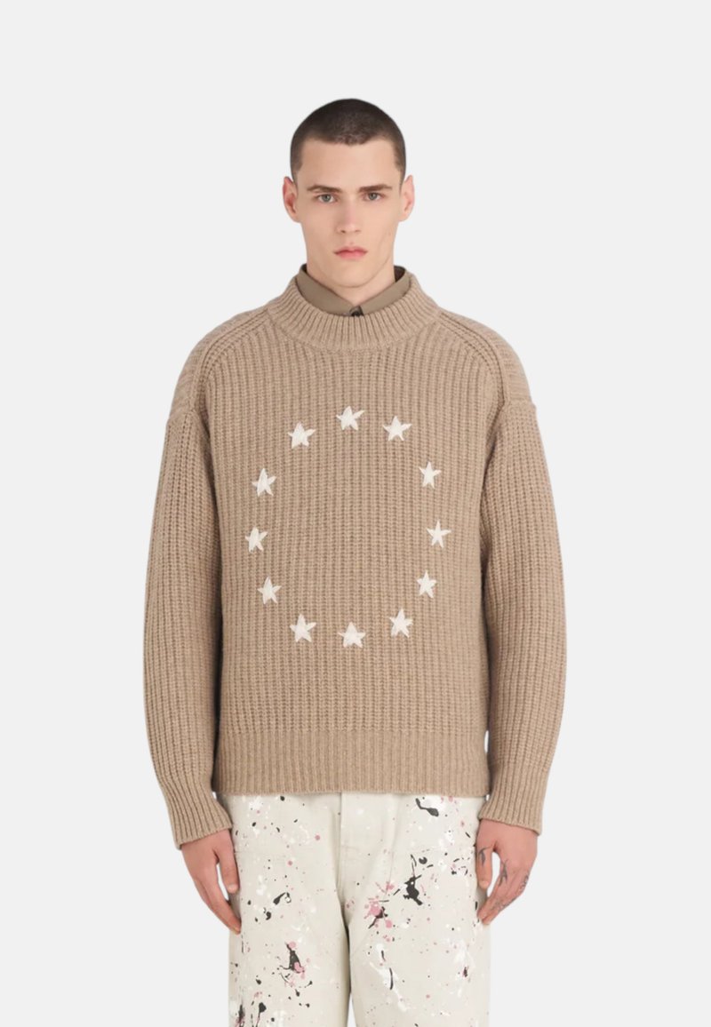 Pull en tricot beige avec un motif circulaire d’étoiles blanches au centre. Présente un col montant et un ourlet côtelé, associé à un pantalon éclaboussé.
