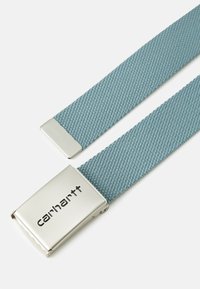 Curea din țesătură împletită de culoare albastru deschis, cu o cataramă metalică argintie care prezintă logo-ul "Carhartt" embosat pe față. Textură netedă, formă rectangulară.