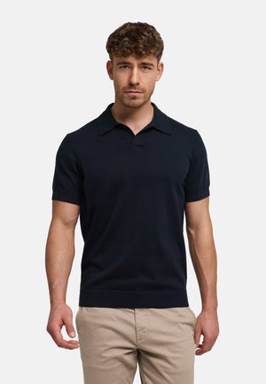 Man met kort krullend haar, gekleed in een zwart poloshirt met korte mouwen en beige broek, staand tegen een effen lichte achtergrond.