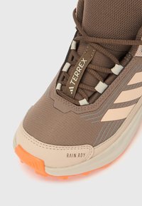 Κοντινό πλάνο καφέ εξω τερικού παπουτσιού Adidas Terrex με λεπτομέρειες μπεζ ρίγας, κορδόνια και πορτοκαλί λαστιχένια σόλα με την ετικέτα RAIN.RDY.
