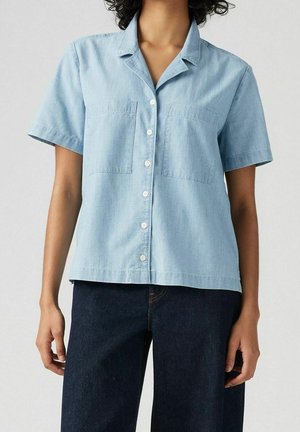 Lyseblå kortærmet skjorte med knapper foran og to brystlommer, båret med mørkeblå bukser på en stående person.