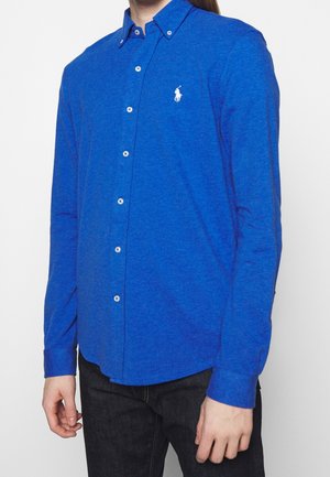 Mann trägt ein langärmeliges, blaues Hemd mit Knöpfen und einem weißen Pferdelogo auf der Brust, kombiniert mit schwarzen Hosen.